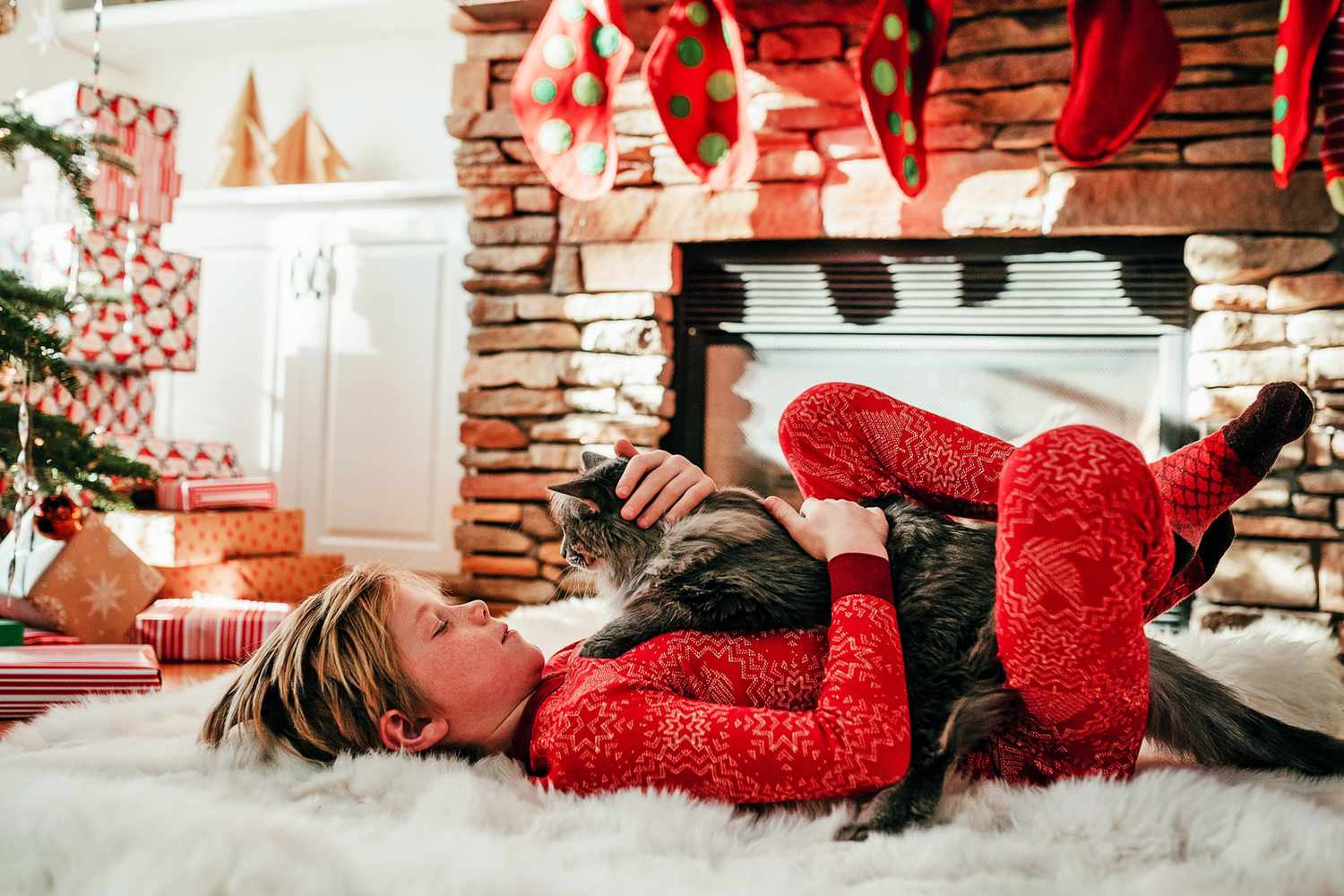 jongen liggend op een wit pluizig tapijt met een kat op zijn borst naast een kerstboom